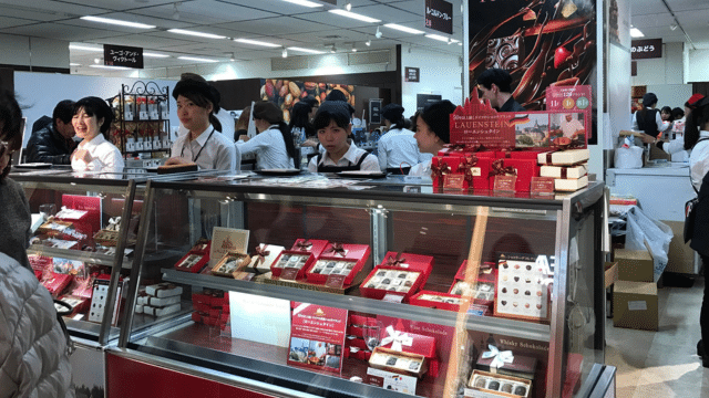 In a kiosk within the store, people in uniforms sell chocolates and other packaged goods, reminiscent of Valentines in Japan where sweets convey heartfelt gestures.