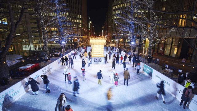 Couples and friends glide gracefully on a brightly lit outdoor rink, surrounded by towering buildings and festive, adorned trees. It's a magical night reminiscent of Valentines in Japan, where romance fills the crisp air beneath the twinkling city lights.