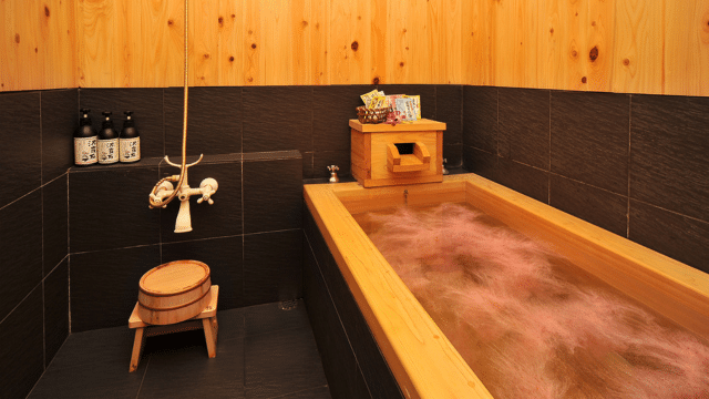 A wooden bathtub filled with water in a room with dark tiles and wooden walls, exuding a serene atmosphere reminiscent of Valentines in Japan, featuring traditional Japanese bath accessories.