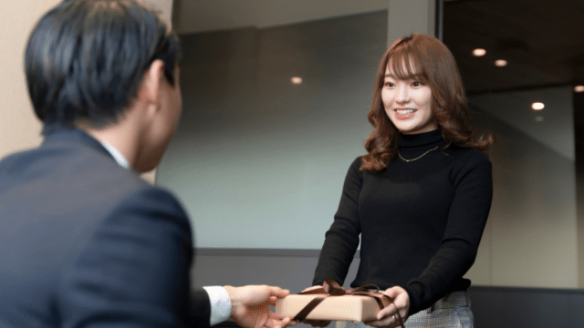 In a Tokyo office, a woman in a black sweater hands a thoughtfully wrapped Valentine's Day gift to a man in a suit, their smiles brightening the professional setting.