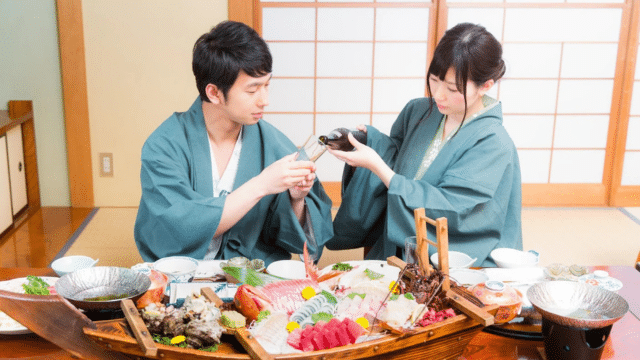 Two people in traditional attire share drinks over a table filled with various sushi and sashimi dishes in a Japanese-style room, capturing the essence of Valentines in Japan.