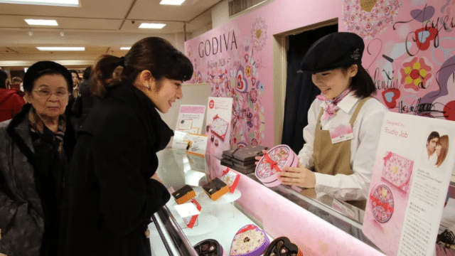 At a Godiva chocolate counter adorned with decorative pink signage, people engage warmly as heart-shaped boxes invite gift-givers. Celebrating Valentines in Japan, this scene captures the essence of sweet connections and shared affection.