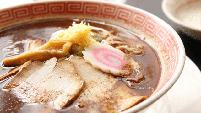 A bowl of ramen with dark broth, topped with sliced pork, bamboo shoots, narutomaki, and shredded green onions, served in a decorative bowl perfect for sharing. Experience a taste of Valentines in Japan with this cozy and delightful meal.