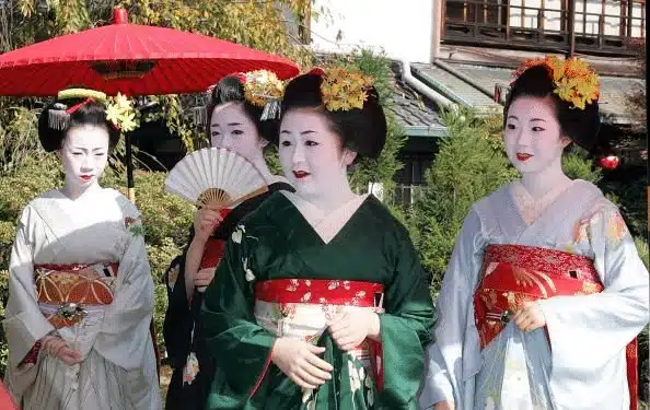 Four women wearing traditional Japanese kimonos and elaborate hairstyles with white face make-up—possibly achieved with a nightingale droppings facial—pose gracefully; one holds a fan, another stands under a red parasol in an outdoor setting.
