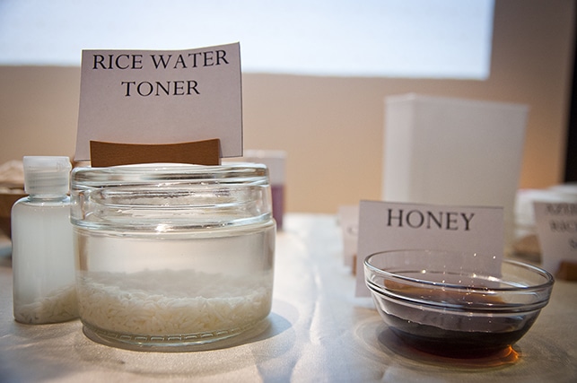 In a beauty workshop, a glass jar labeled "Rice Water Toner" containing liquid with rice grains sits next to a bowl labeled "Honey" on a table with other labeled containers in the background.