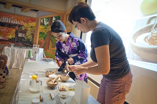 Two people prepare ingredients on a table, one wearing a floral kimono, the other in a T-shirt. The table holds various bowls and utensils, suggesting this might be part of a beauty workshop. A presentation is visible in the background.