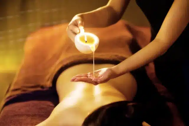 A person pours oil onto their hand above the back of someone lying face down on a massage table in a dimly lit room, creating a soothing setting for a candle massage.