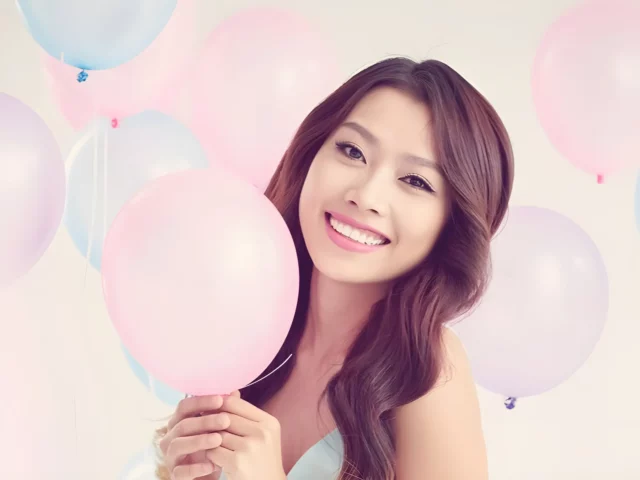 A woman with long brown hair smiles while holding a pink birthday balloon, surrounded by pastel-colored balloons against a light background.