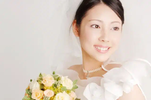 A radiant bride, glowing from a recent bride spa, wears a white dress and veil as she smiles warmly, holding a bouquet of yellow and white roses and looking to the side.