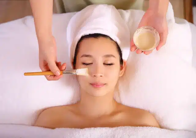 Person lying on a spa bed with a towel wrapped around their head, receiving an organic facial treatment as an aesthetician applies a mask with a brush.