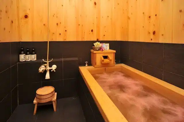 A Japanese-style bathroom with a wooden soaking tub filled with water, black tile walls and floor, a wooden stool, and bath products on the ledge.