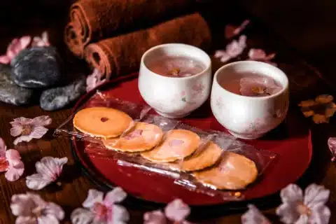 Two cups of tea with cherry blossom petals, packaged cookies, towels, and decorative stones arranged on a tray with scattered pink flower petals.