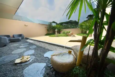 A Japanese-style zen garden with gravel paths, a stone basin with a bamboo ladle, stone lantern, greenery, and modern outdoor seating. Sandals are placed on the gravel.