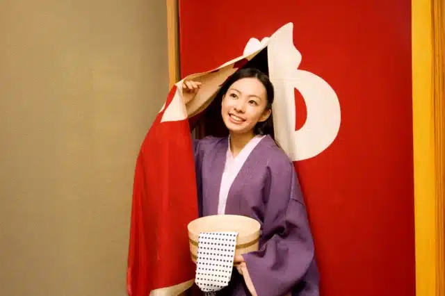 A woman in a purple kimono holds a wooden bucket and lifts a red curtain with a white symbol, standing at the entrance of a Japanese bathhouse—a serene scene that makes a unique gift for girlfriend.