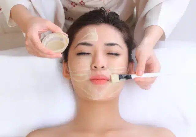 A person lying down is having an organic facial mask applied with a brush by another person.