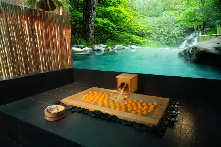 A wooden indoor bath filled with whole oranges next to a flowing onsen pool, surrounded by greenery and a bamboo screen.