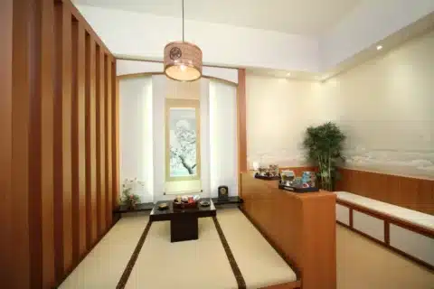 A minimalist Japanese-style tatami room with a low table, cushions, wall scroll, decorative plants, and soft lighting.