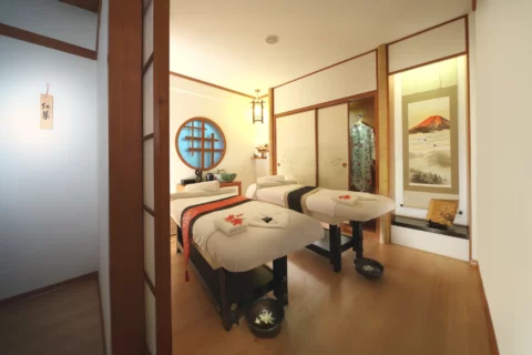 A Japanese-style spa room with two massage tables, wall art, soft lighting, and traditional decor elements, creating a calm and serene atmosphere.