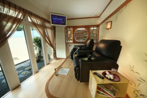A cozy room with two black recliner chairs, a mounted TV, wall decor, sheer curtains, and a small wooden table with books, a remote, and a drink.