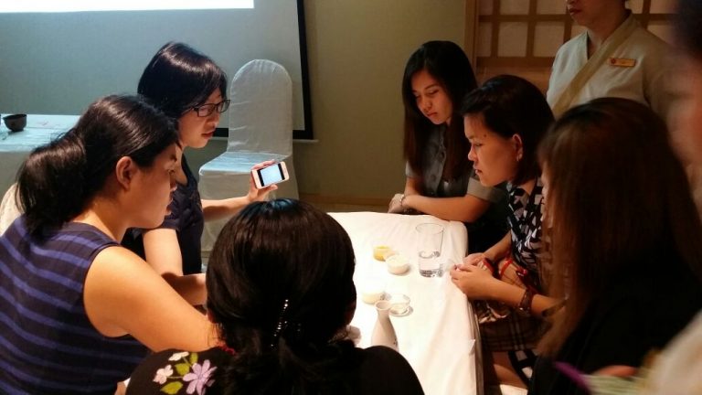 A group of women sitting around a table, looking intently at a phone held by one of them. Cups and glasses are scattered on the table, hinting at discussions from an engaging beauty event. A presentation screen is partially visible in the background.