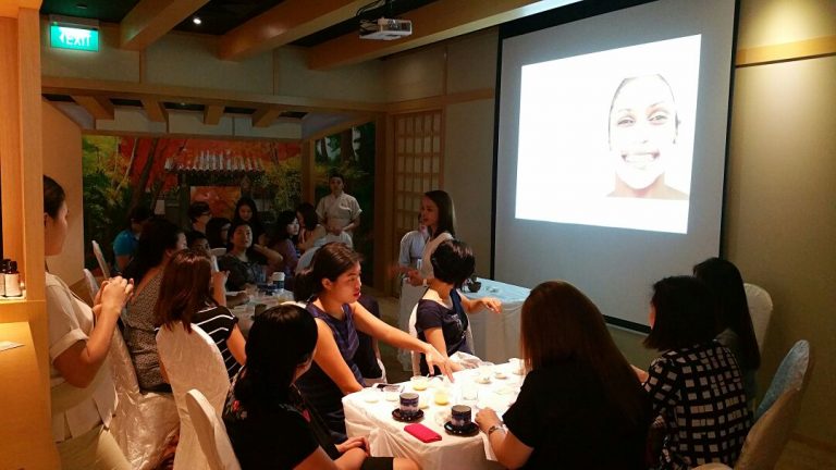 A group of people are seated around a table in a meeting room, attentively listening to a speaker standing near a projection screen showing a smiling face, as they discuss details for an upcoming beauty event.