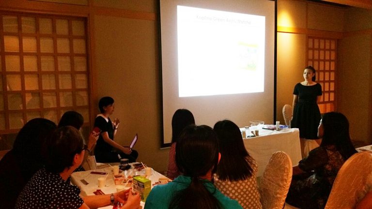 A group of people seated around a table watch a presentation on a screen in a dimly lit room, setting the scene for an exclusive beauty event. One person is presenting while another sits next to the projector screen.