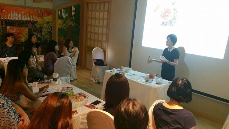 A person stands and gives a presentation to a group seated around a table at a beauty event, with a laptop and various objects on the table, and a large screen displaying an image in the background.