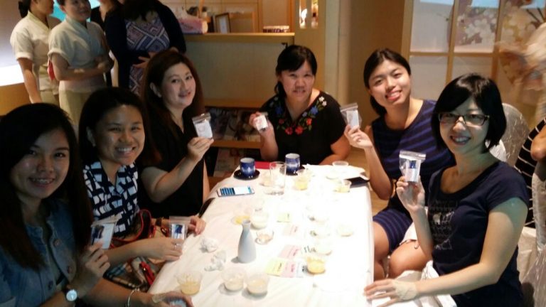 A group of six smiling individuals sits around a table at a beauty event, holding small packages. The table is covered with various items including cups, plates, and papers.