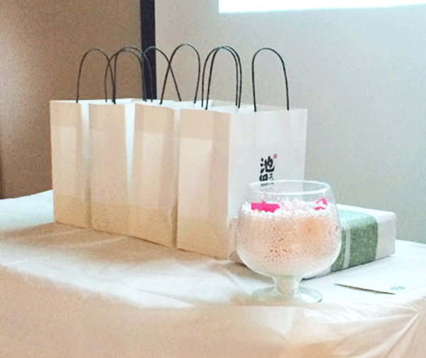 A table with gift bags, a glass bowl filled with small white balls, and a wrapped gift box sets the scene for a delightful beauty event.