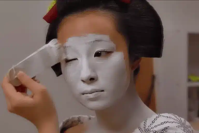 A person applies white makeup to their face with a brush, possibly preparing for a traditional Japanese geisha performance.