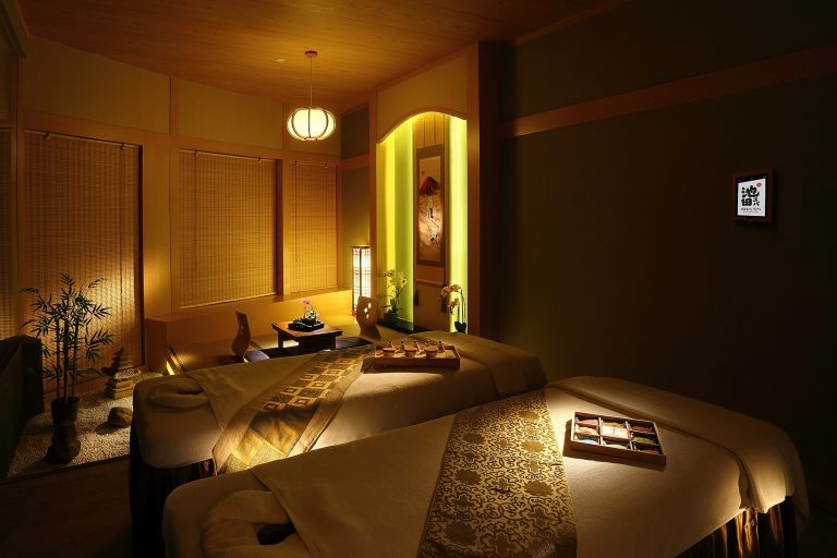 A warmly lit spa room with two massage tables adorned with towels and decor, perfect for a beauty event. Walls are decorated with soft lighting, bamboo elements, and a small Japanese art piece.