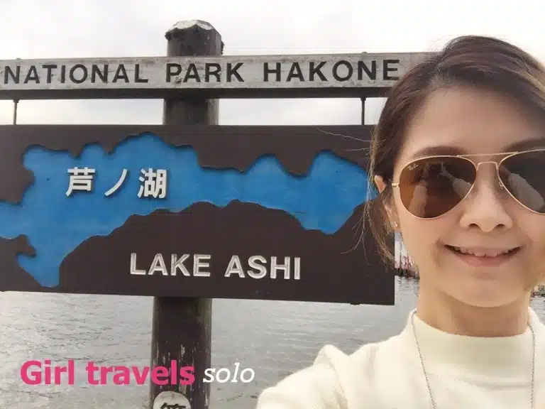A woman wearing sunglasses takes a selfie in front of a Lake Ashi sign at Hakone National Park. Text on the photo reads, "Girl travels solo—Tokyo solo trip adventure.