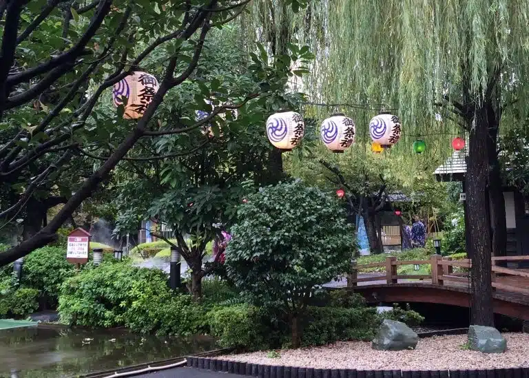 Japanese paper lanterns hang from tree branches above a wooden bridge in a lush garden—an inviting scene for anyone enjoying a peaceful Tokyo solo holiday surrounded by vibrant greenery.