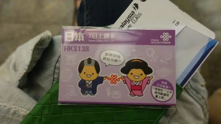 A Japan 7-day data SIM card package with cartoon characters is held over a green woven bag and a boarding pass—perfect for your Tokyo solo holiday.
