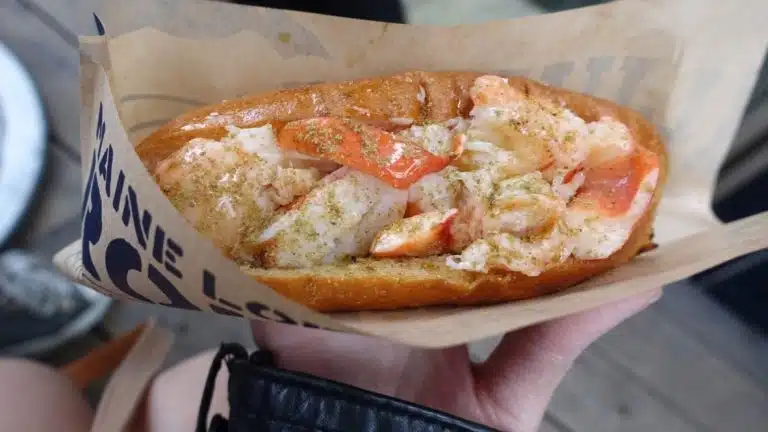 A hand holds a lobster roll filled with chunks of lobster meat and sliced red pepper in a toasted bun, wrapped in brown paper—a perfect treat to savour on your Tokyo solo trip.