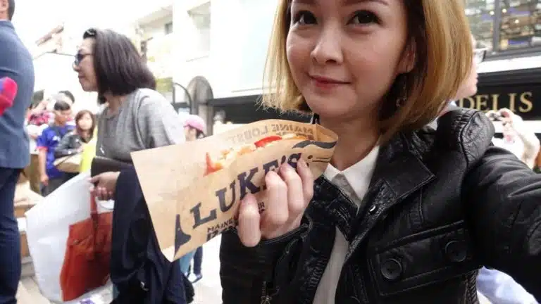 A woman in a black jacket holds a Luke's sandwich wrapper whilst standing outdoors among a crowd of people during her Tokyo solo trip.