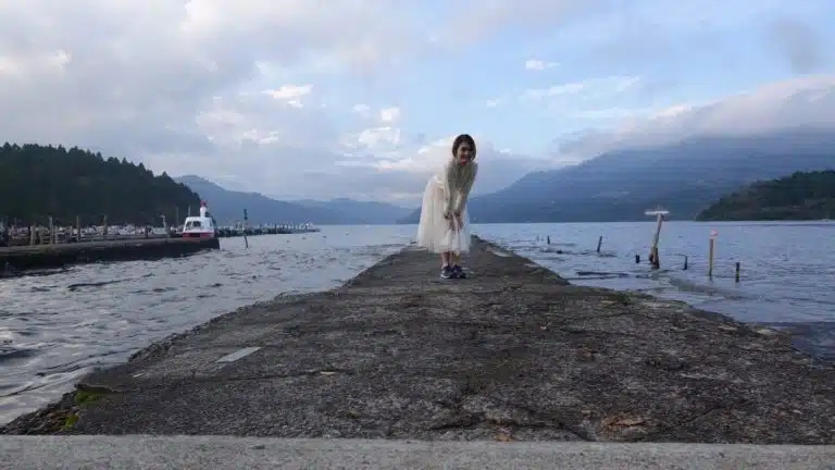 A person on a Tokyo solo trip in a white outfit stands on a concrete pier extending into a lake, with mountains and a moored boat in the background under a cloudy sky.
