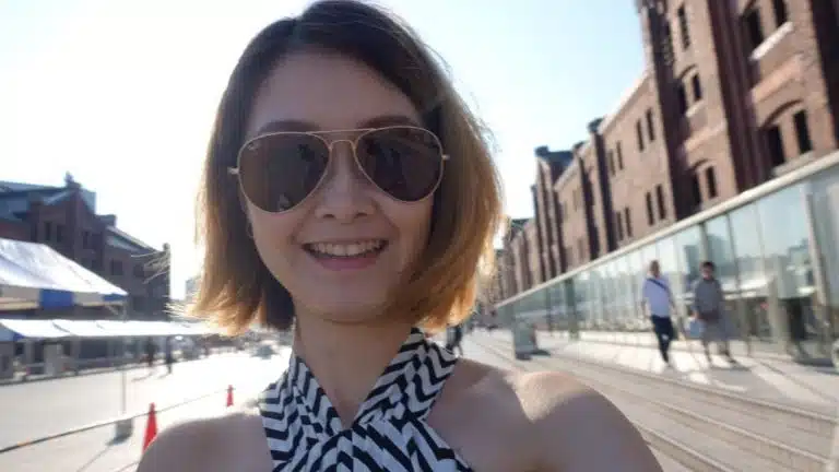 A woman on a Tokyo solo trip, wearing sunglasses and a striped halterneck top, smiles at the camera outdoors, with brick buildings and a glass balustrade in the background.
