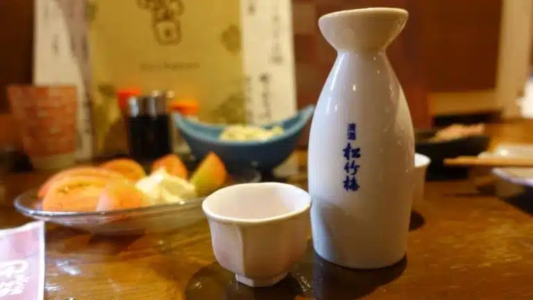 A ceramic sake bottle and cup are placed on a wooden table with sliced tomatoes and other dishes in the background—capturing the essence of a Tokyo solo trip meal.
