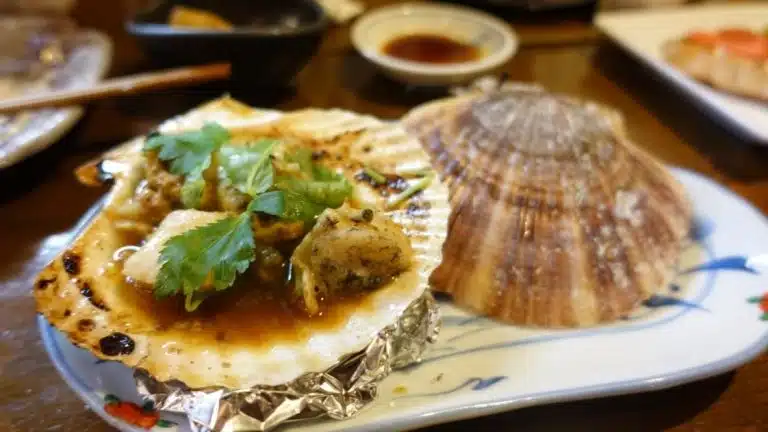 A cooked scallop in its shell with sauce and coriander garnish, next to an empty shell on a plate—perfect for savouring during a Tokyo solo trip, with other dishes in the blurred background.