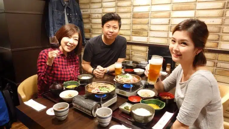 Three people sitting at a restaurant table in Tokyo with various dishes and drinks, smiling at the camera—capturing the joy of friendship during a Tokyo solo holiday.