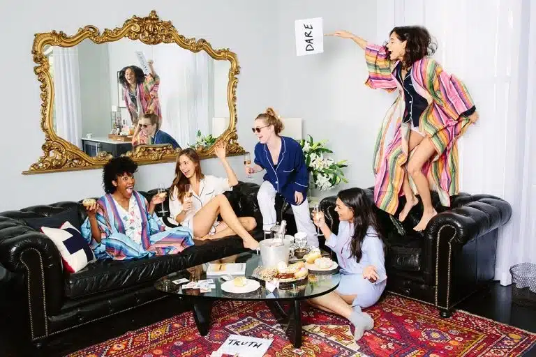 Five women in pyjamas play a game of Truth or Dare in a lounge, one holding a "DARE" sign while standing on the sofa; snacks and drinks fill the glass coffee table during their fun Valentines 2017 celebration.