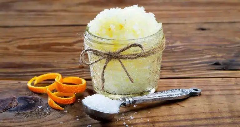 A glass jar filled with coarse, yellowish body scrub tied with string sits next to orange peel and a metal spoon with salt on a wooden surface.
