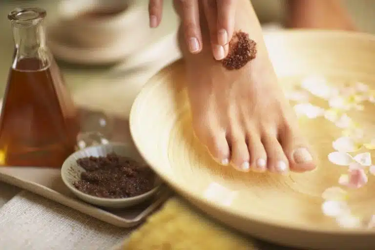 A person applies a brown body scrub to their foot over a bowl of water with flower petals; a jar of oil and a dish of scrub are nearby.
