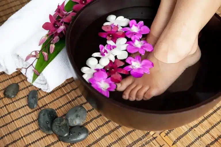 A person's feet soaking in a bowl of water with floating pink and white flowers, next to a towel, orchids, smooth black stones, and a body scrub on a bamboo mat.