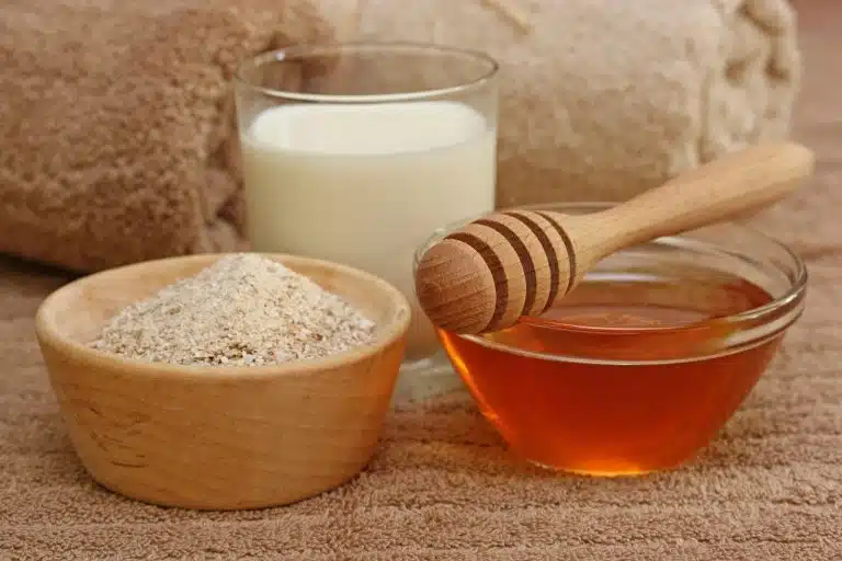 A bowl of porridge, a glass of milk, and a bowl of honey with a wooden drizzler are arranged on a brown towel, ready to become the perfect ingredients for a nourishing homemade body scrub.
