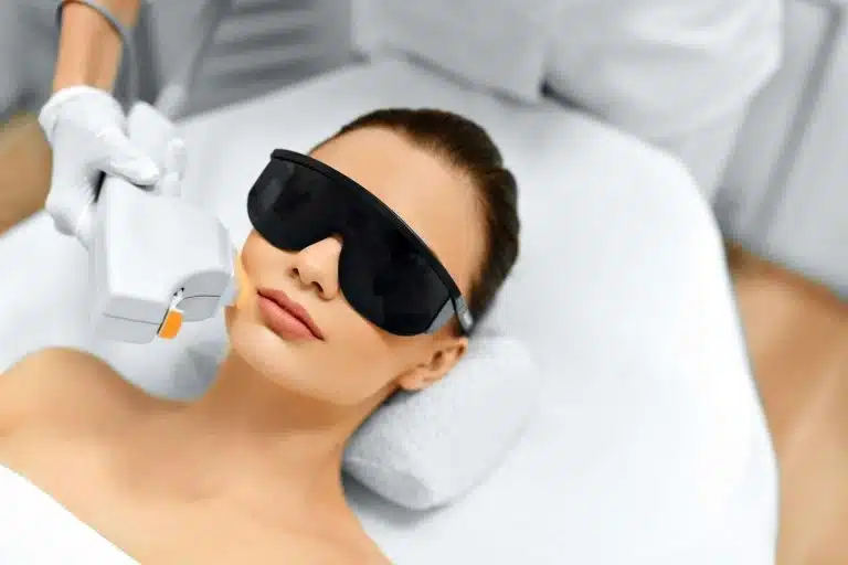 A person lying on a treatment bed wears protective goggles whilst receiving a facial care procedure with a handheld laser device on the face.