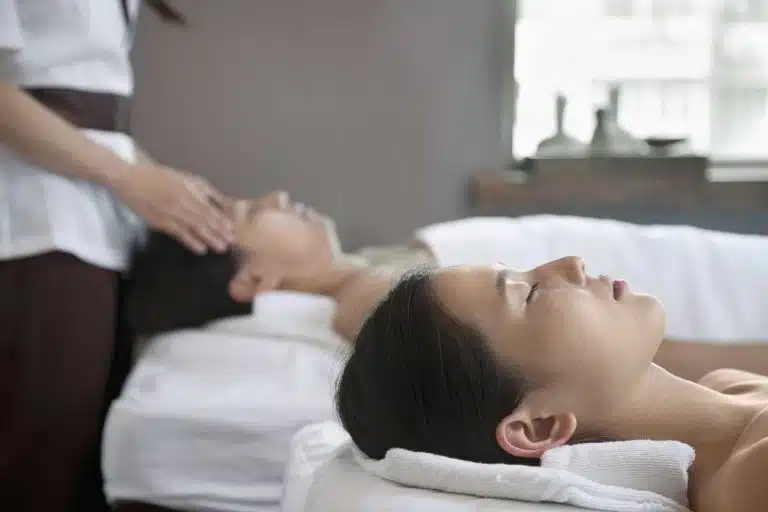 Two people lying on massage tables with eyes closed; one enjoys a soothing head massage from a therapist while the other relaxes beside them—a perfect Mother’s Day gift experience in a serene spa setting.