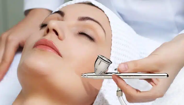 A person with a towel wrapped around their head receives facial care as a practitioner uses an airbrush device for the treatment.