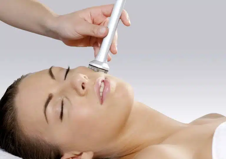 A person enjoys facial care, receiving a treatment with a handheld device whilst lying down with eyes closed.